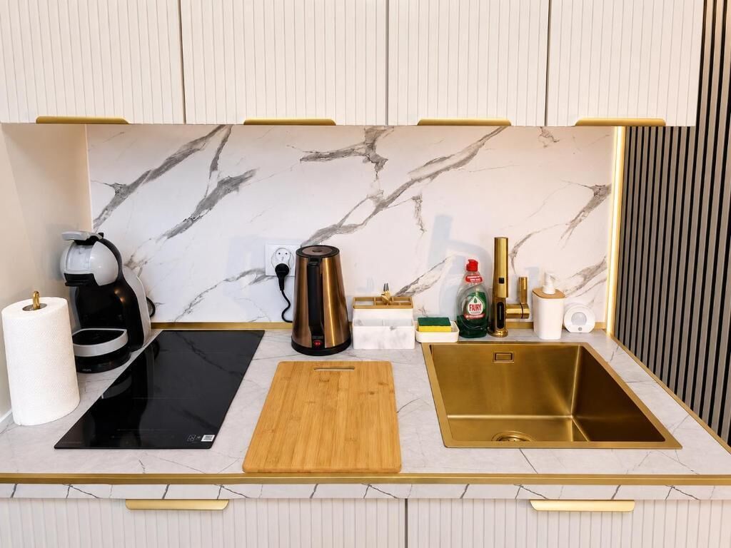 Una cocina moderna con salpicadero de mármol, placa de inducción negra, tabla de cortar de madera, pileta dorada y diversos electrodomésticos de cocina.