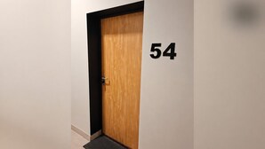 A wooden door with a black frame and a black doormat on a tiled floor. The number '54' is visible on the wall to the right of the door.