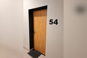 A wooden door with a black frame and a black doormat on a tiled floor. The number '54' is visible on the wall to the right of the door.