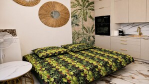 Quarto com cama, ventilador, espelho redondo, painel na parede e área de cozinha com armários e pia.