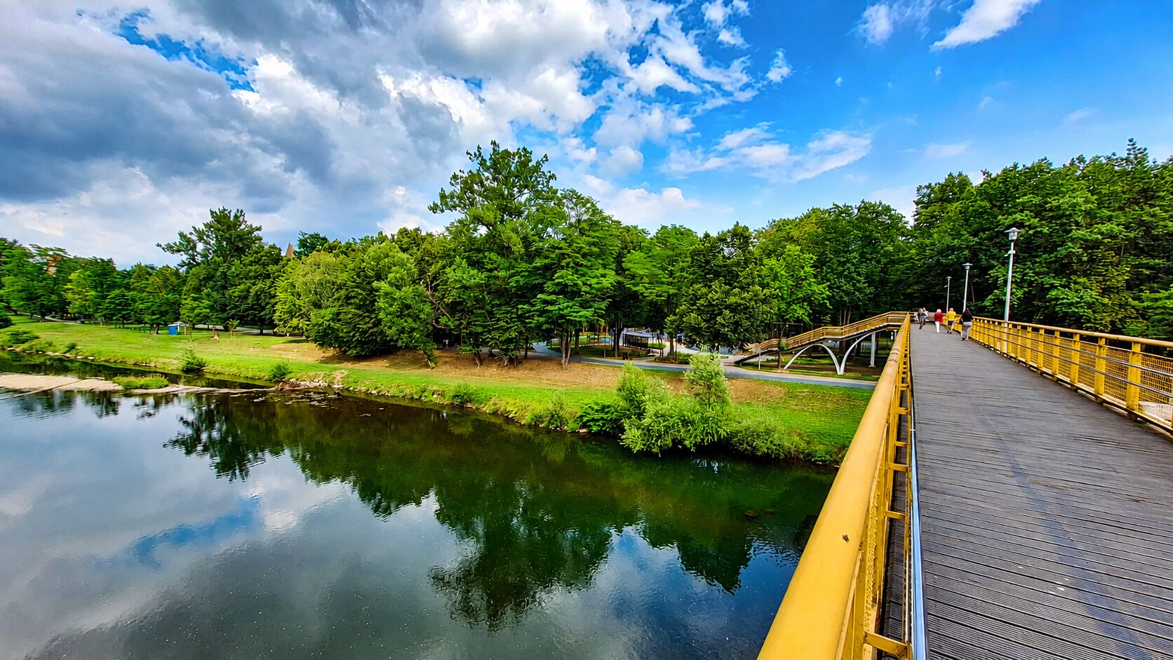 A wooden bridge over a calm river with a clear sky and lush greenery.