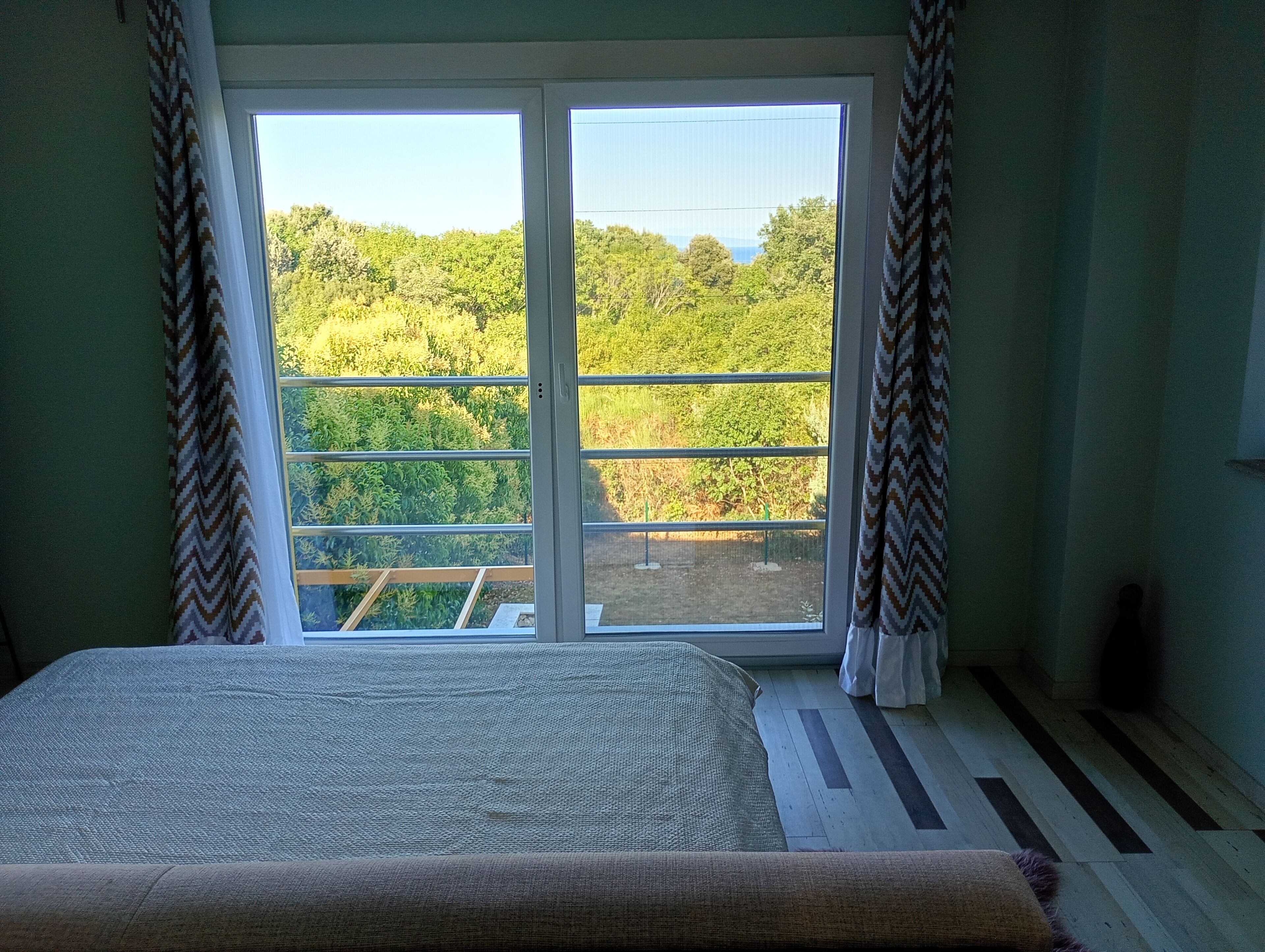 A room with a bed, a view of greenery through a window, and a patterned curtain.