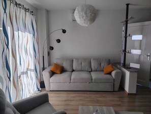 Un salon moderne avec un canapé gris, deux coussins orange, un lampadaire noir et une suspension blanche.