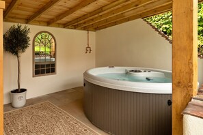 Ein Whirlpool unter einer Holzpergola mit einem Topfbaum und einem Fenster mit Blick auf einen Garten.