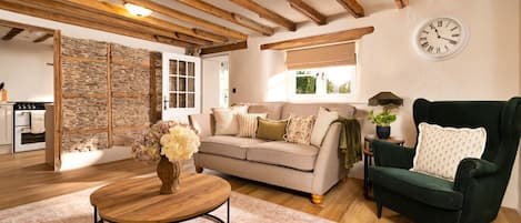Un salon chaleureux avec un plafond en bois, un canapé, un fauteuil vert, une table basse ronde et une horloge au mur.