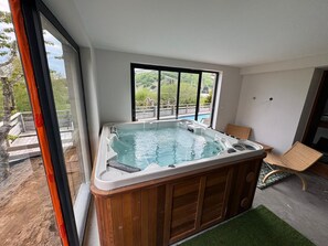 A hot tub in a room with a large window, wooden flooring, and a chair. - Chalet for rent with a south-facing heated outdoor pool (Besse-et-Saint-Anastaise)