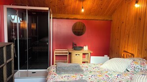 Een slaapkamer met een houten plafond, een bed met bloemenbeddengoed, een nachtkastje met een schaal en flessen, en een schuifdeur naar een doucheruimte.