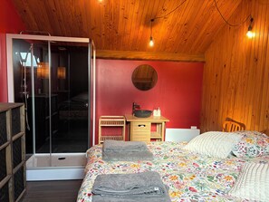 Quarto com teto de madeira, cama com roupa de cama floral, mesa de cabeceira com uma tigela e garrafas, e porta de vidro deslizante para área do chuveiro.