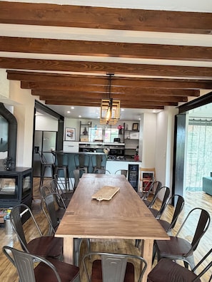 Área de refeições com mesa de madeira e cadeiras de metal, cozinha ao fundo e uma janela grande.