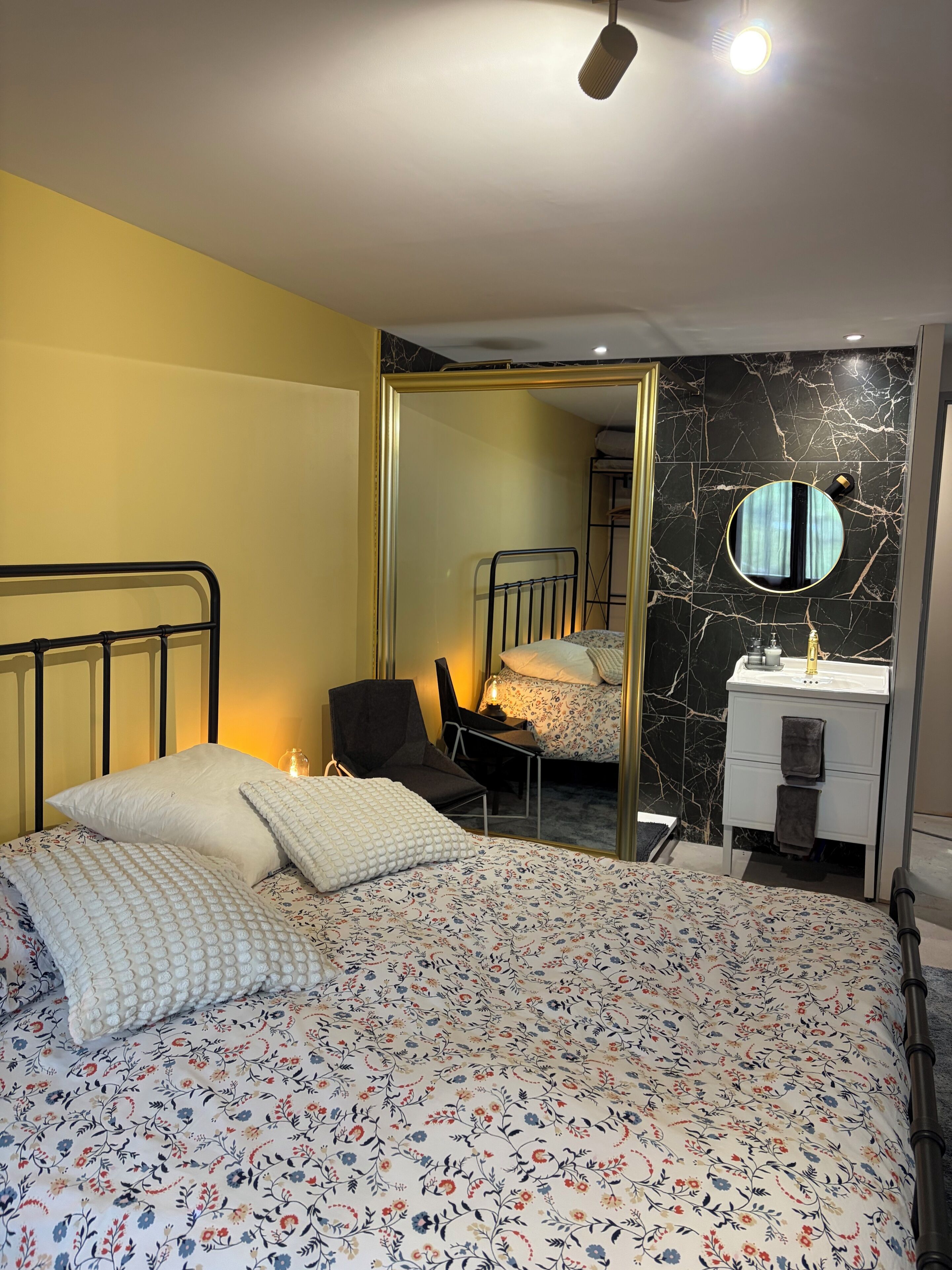 A bedroom with a bed, a chair, a bedside table, a mirror, and a bathroom with a sink and mirror.