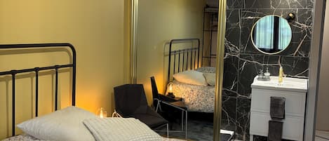 A bedroom with a bed, a chair, a bedside table, a mirror, and a bathroom with a sink and mirror.