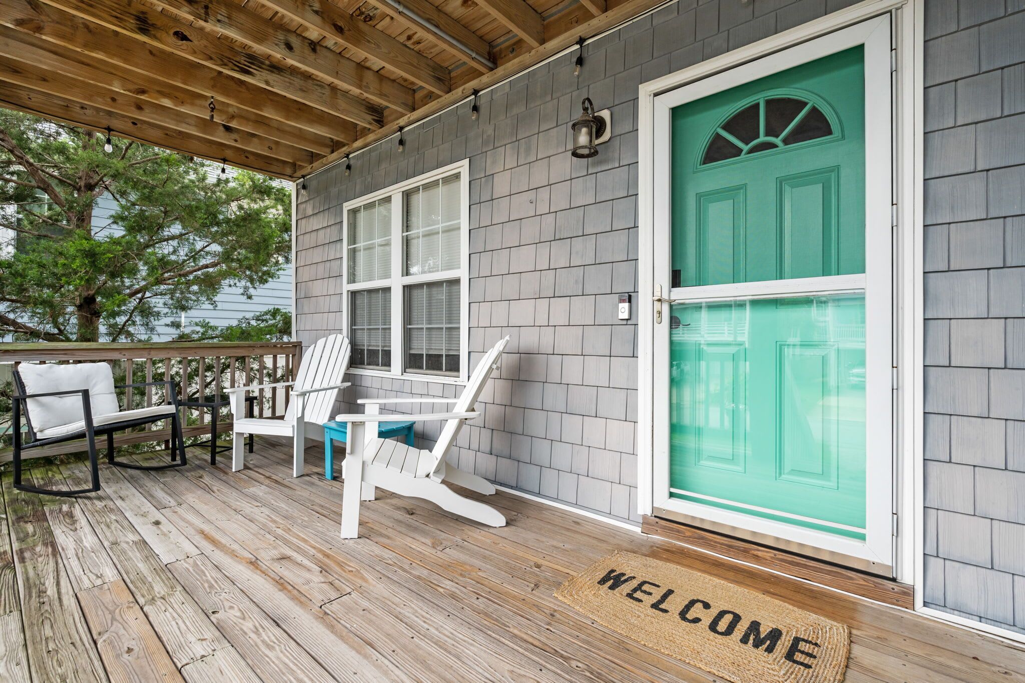 A covered porch with a wooden deck, a white chair, a blue chair, and a green door with a welcome mat.