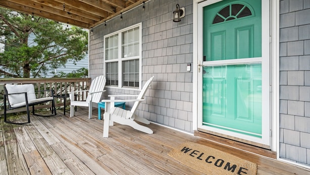 A covered porch with a wooden deck, a white chair, a blue chair, and a green door with a welcome mat. - Coco Beach - Walk to Carolina Beach, Family Friendly Townhome (Carolina Beach)