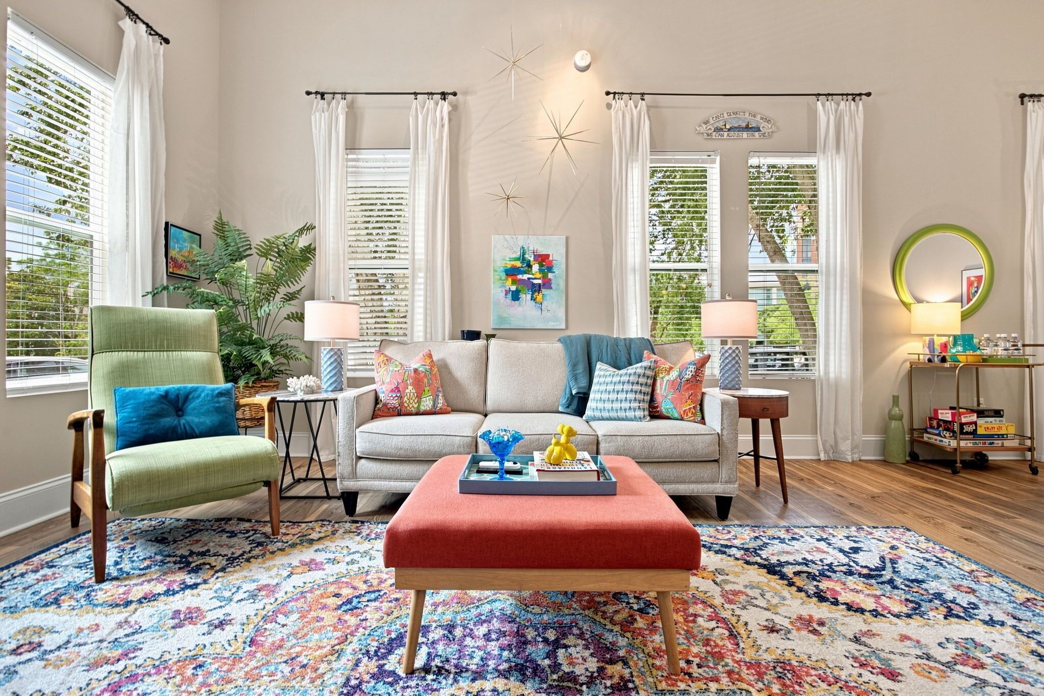 A living room with a sofa, armchairs, a coffee table, and a rug. There are windows with curtains, a wall clock, and a decorative star on the wall.