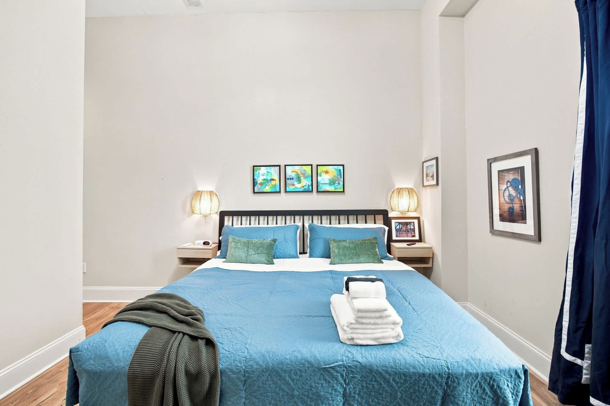 A neatly made bed with blue bedding, two bedside lamps, and a stack of towels.