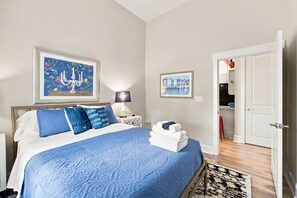 A bedroom with a bed, pillows, a nightstand, a lamp, a framed picture, a rolled towel, and a door leading to a kitchen. - Brooklyn Arts Condo | Walk to Everything (Wilmington)