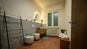 Een moderne badkamer met een stenen muur, een houten wastafelmeubel, twee toiletten, een bidet, een spiegel met verlichting en een raam met gezandstraald glas.