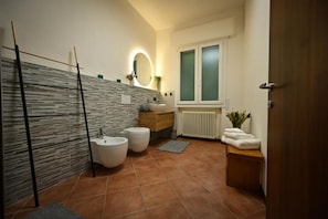 Ein modernes Badezimmer mit Steinwand, Holzwaschtisch, zwei Toiletten, einem Bidet, einem Spiegel mit Beleuchtung und einem Fenster mit Milchglas.