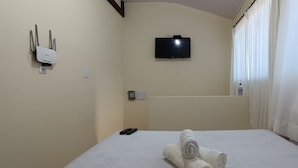 A hotel room with a bed, a TV mounted on the wall, a power outlet, and a bottle of hand sanitizer. - Apto Econômico Mafra  (Mafra)