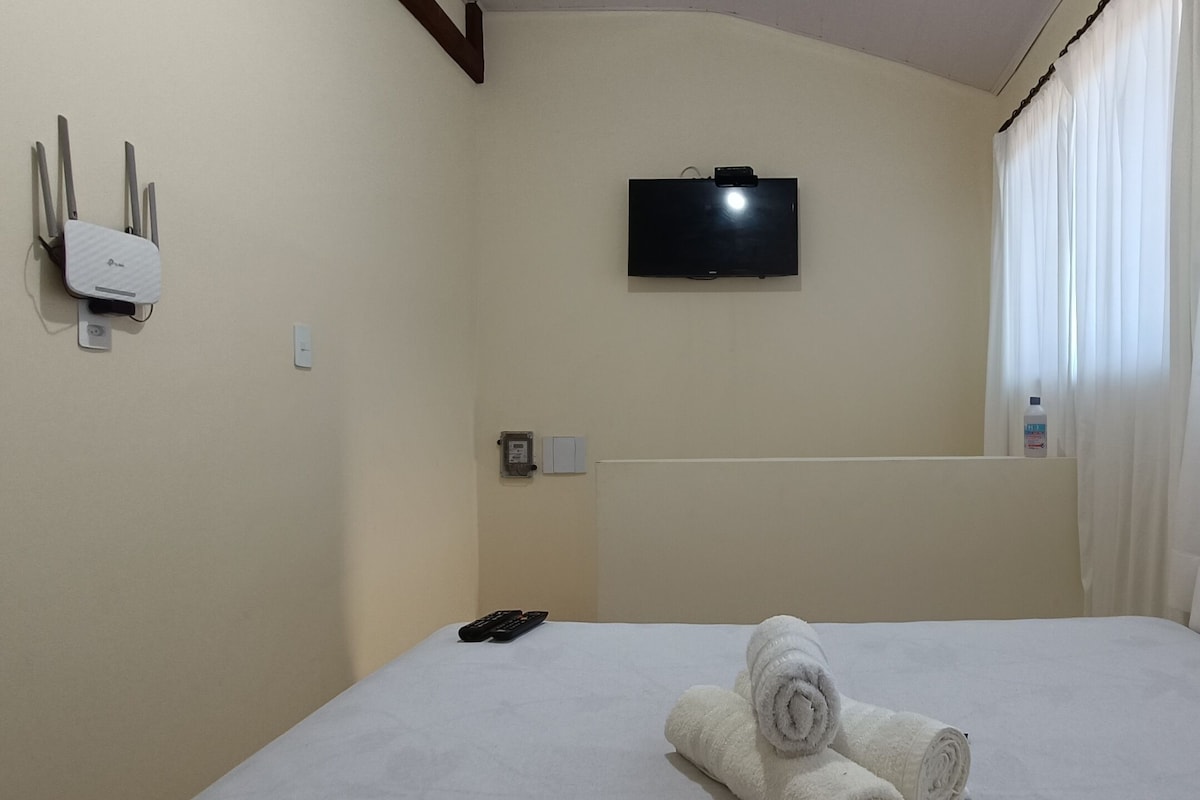 A hotel room with a bed, a TV mounted on the wall, a power outlet, and a bottle of hand sanitizer.
