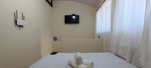 A hotel room with a bed, a TV mounted on the wall, a power outlet, and a bottle of hand sanitizer. - Apto Econômico Mafra  (Mafra)