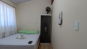A small, single-bed room with a green pillow, a rolled towel, and two remote controls on the bed. There is a black wardrobe with a fan on top, a light switch, and a power outlet on the wall. - Apto Econômico Mafra  (Mafra)