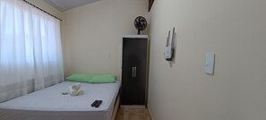 A small, single-bed room with a green pillow, a rolled towel, and two remote controls on the bed. There is a black wardrobe with a fan on top, a light switch, and a power outlet on the wall. - Apto Econômico Mafra  (Mafra)