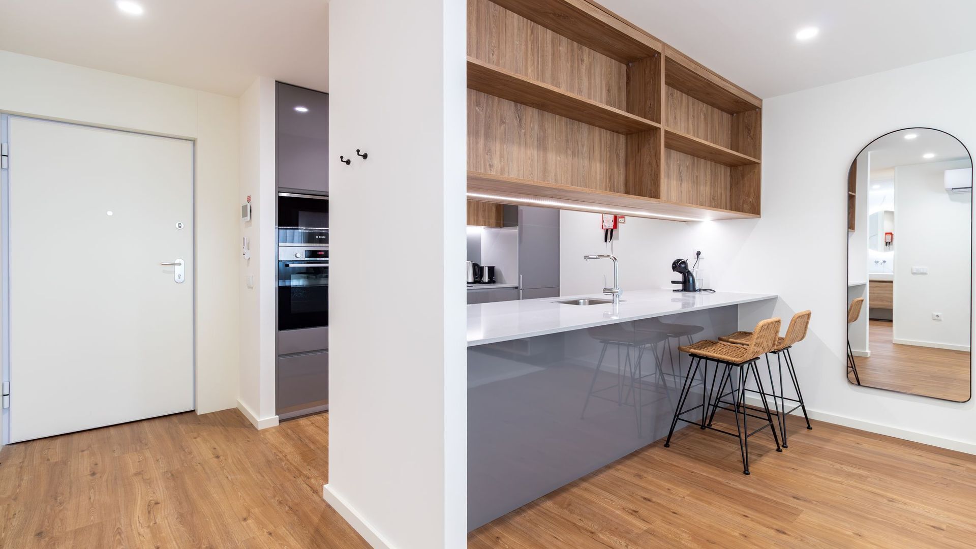 Une cuisine moderne dotée d’un four encastré, d’un plan de travail blanc et d’étagères en bois. On y trouve deux tabourets de bar et un grand miroir accroché au mur.