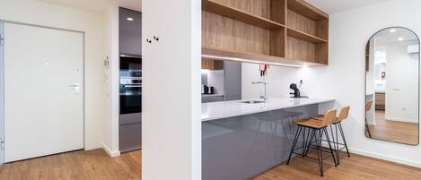 Une cuisine moderne dotée d’un four encastré, d’un plan de travail blanc et d’étagères en bois. On y trouve deux tabourets de bar et un grand miroir accroché au mur.