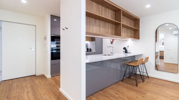 Eine moderne Küche mit Einbauofen, weißer Arbeitsplatte und Holzregalen. Zwei Barhocker und ein großes Spiegelbild an der Wand sind ebenfalls vorhanden.