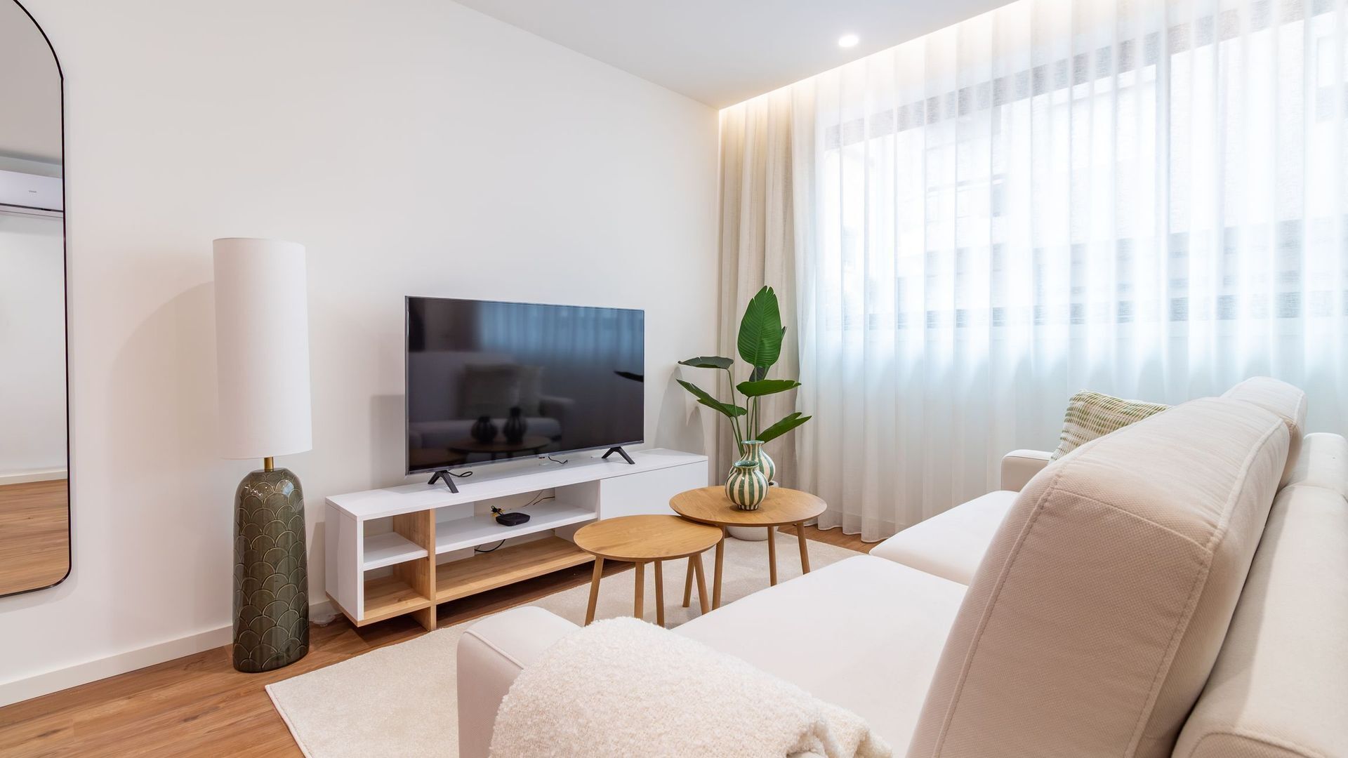 Uma sala de estar moderna com televisão de tela plana, um sofá branco, uma mesa de madeira pequena e um vaso com planta.