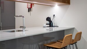 Une cuisine moderne avec un plan de travail blanc, un évier en acier inoxydable et une machine à café. On y trouve deux chaises en osier et une petite table avec une tasse et une soucoupe.
