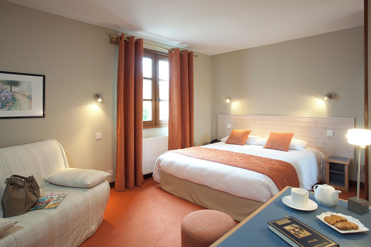 Een hotelkamer met een bed, een stoel, een tafel met een kopje en koekjes, en een raam met gordijnen.