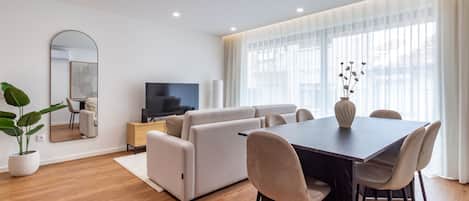Un salon moderne comprenant un ensemble de canapés, une table à manger avec des chaises, un téléviseur à écran plat et une grande fenêtre ornée de rideaux légers.