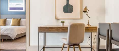 A bedroom with a bed, a desk with a chair, a framed picture, and a small plant.