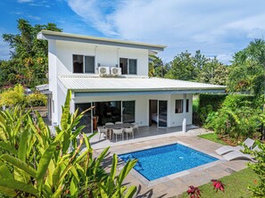 Una casa moderna de dos pisos con piscina, mobiliario exterior y abundante vegetación.