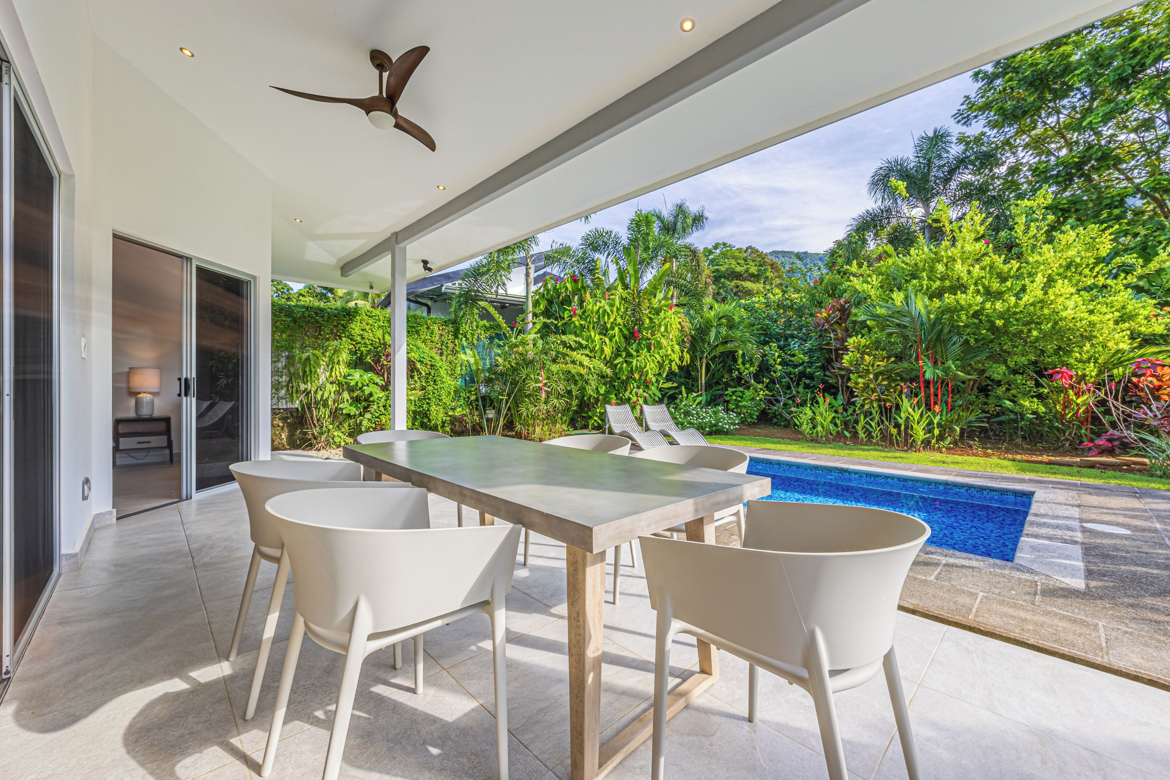Un patio exterior con una mesa y sillas de comedor, un ventilador de techo y vistas a una piscina rodeada de abundante vegetación.