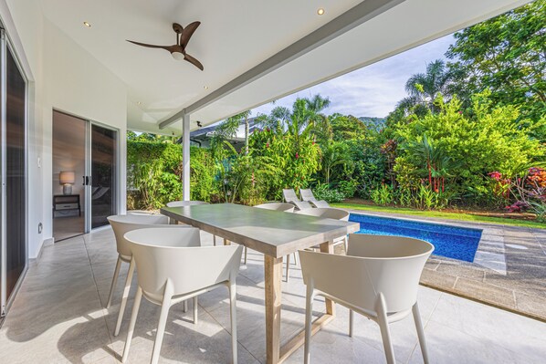 Un patio exterior con una mesa y sillas de comedor, un ventilador de techo y vistas a una piscina rodeada de abundante vegetación.