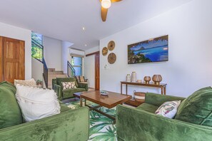 Amplia sala de estar con escalera, mobiliario tapizado en verde, una mesa de centro de madera y un cuadro colgado en la pared.