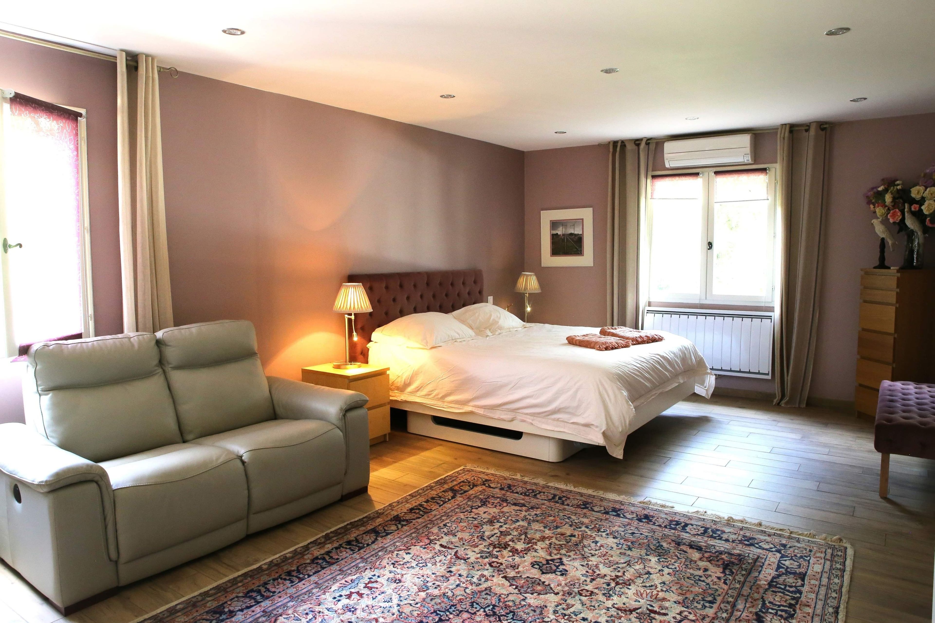 Une chambre à coucher comprenant un lit, une chaise, une table de chevet, une lampe, une fenêtre avec des rideaux et un radiateur.