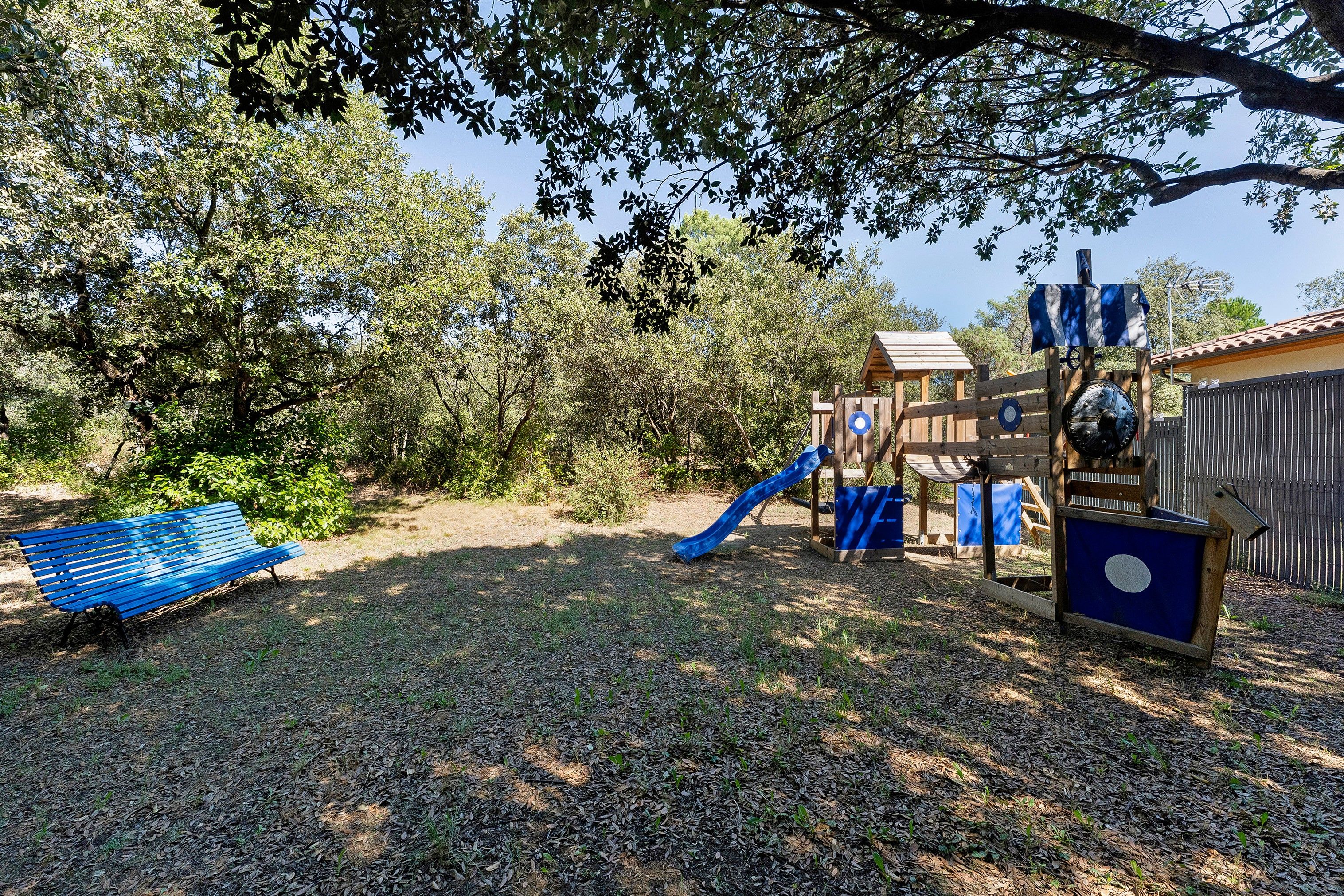 Un parque infantil con un tobogán y estructuras para escalar, rodeado de árboles y un banco.