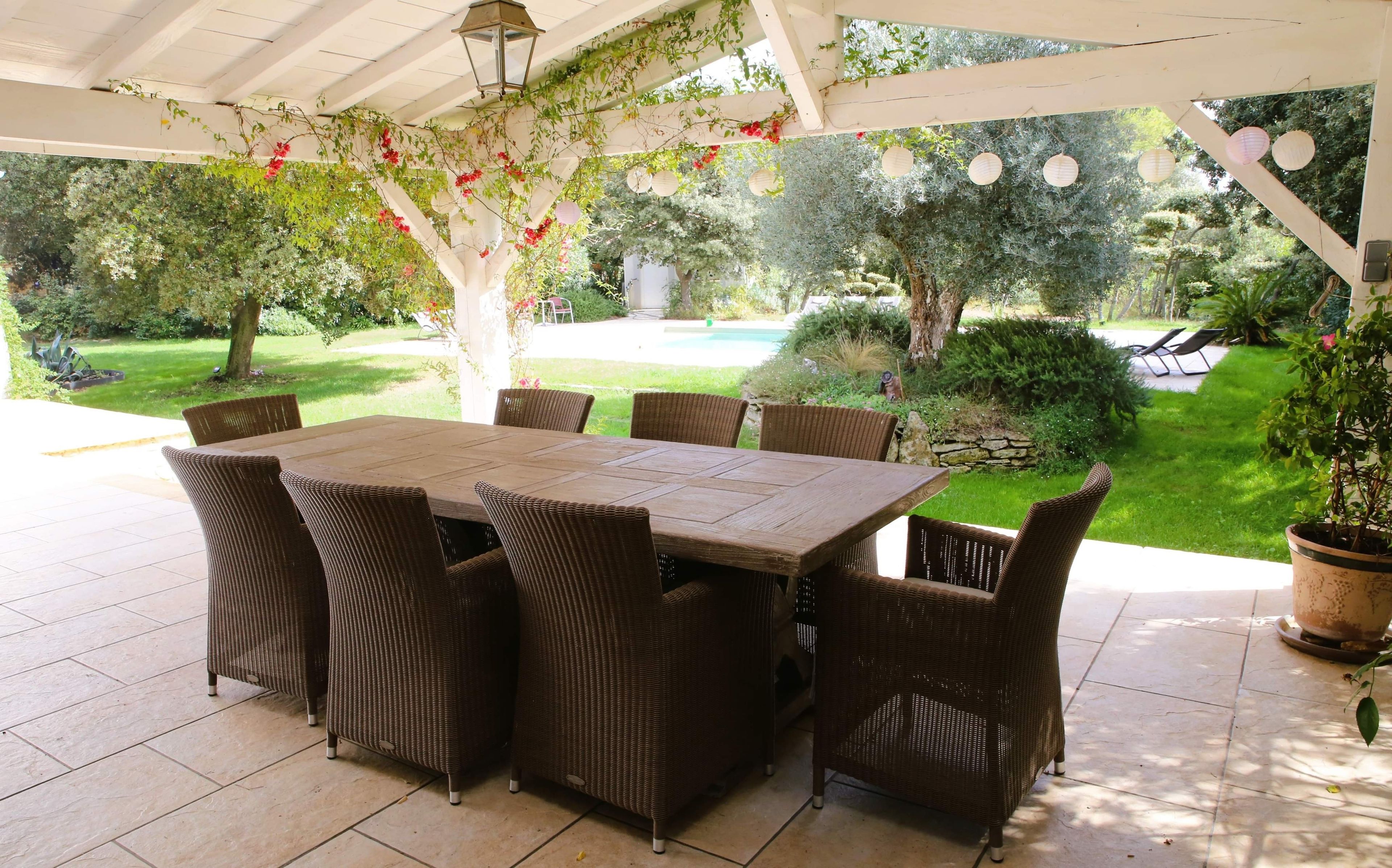 A covered patio with a table and chairs, surrounded by greenery and a pool.