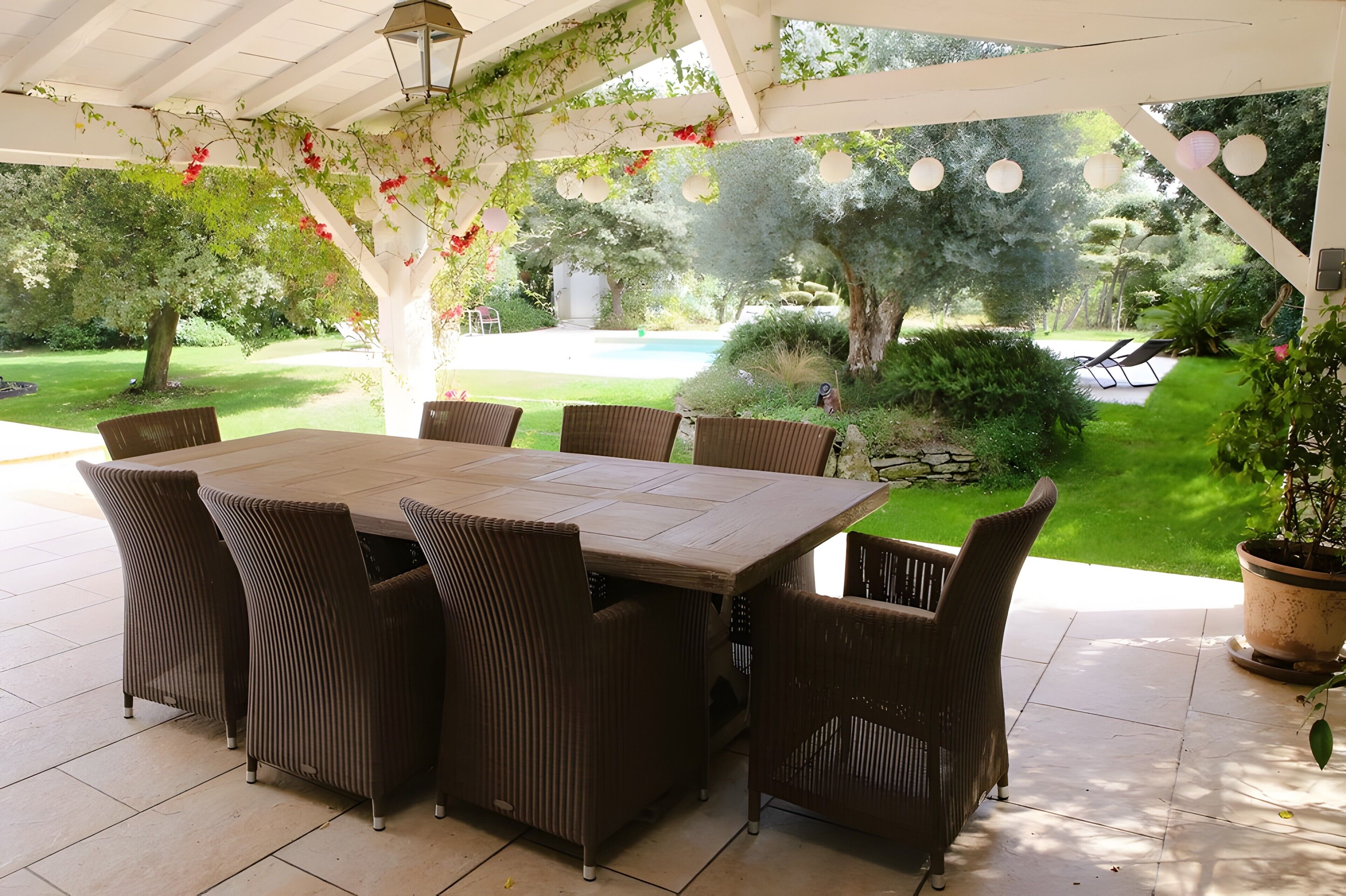 Une terrasse couverte avec une table et des chaises en bois, entourée de verdure et offrant une vue sur une piscine.