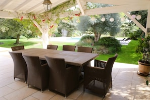 Une terrasse couverte avec une table et des chaises en bois, entourée de verdure et offrant une vue sur une piscine.