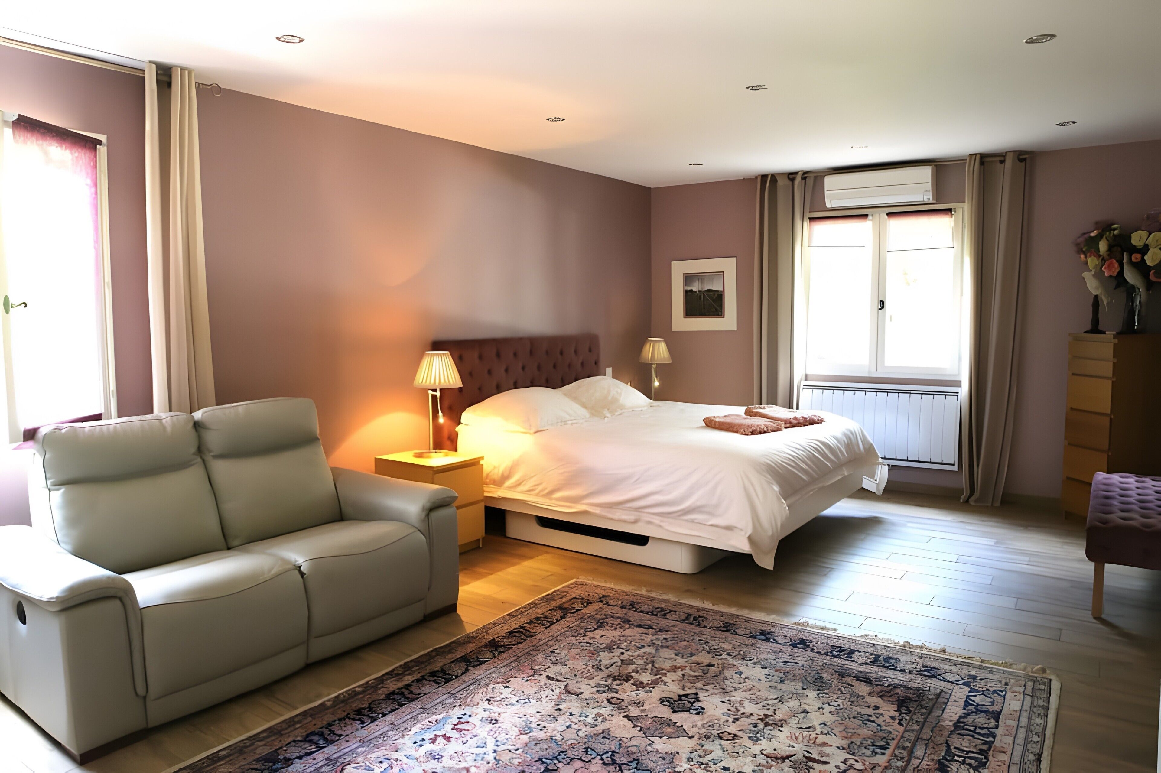 Une chambre à coucher comprenant un lit, une chaise, une table de chevet, une lampe, une fenêtre avec des rideaux et un radiateur.