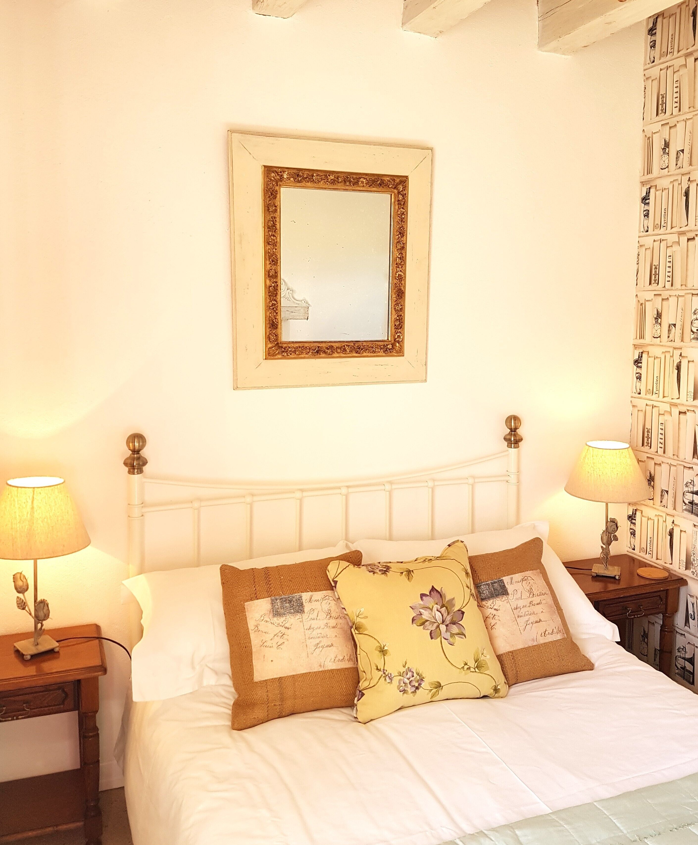 A bedroom with a bed, two bedside lamps, a nightstand, a mirror, and a wall with a bookshelf.
