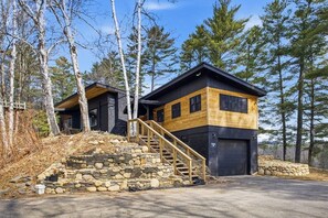 Une cabane moderne avec un escalier en pierre menant à une terrasse en bois, le tout entouré d’arbres.