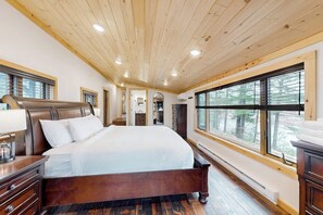 Un dormitorio con techo de madera, ventanas amplias, cama, mesita de noche y cómoda.