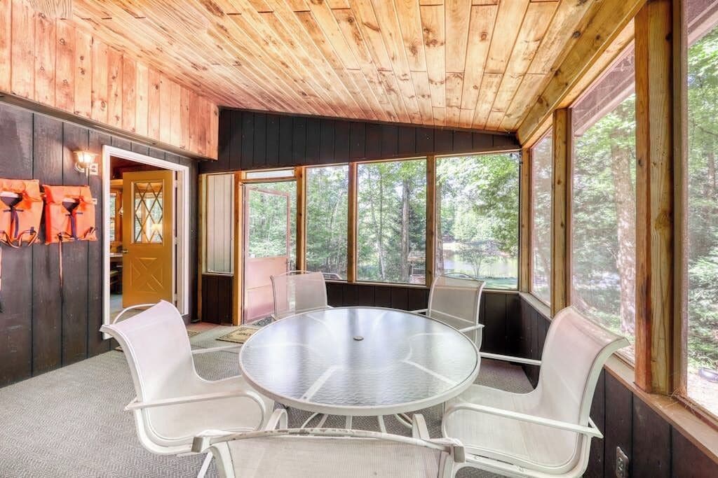 A room with a round glass table surrounded by white chairs, large windows, and wooden walls.