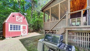 Una stalla rossa e una casa di legno con veranda, immerse in un prato erboso e circondate da alberi.